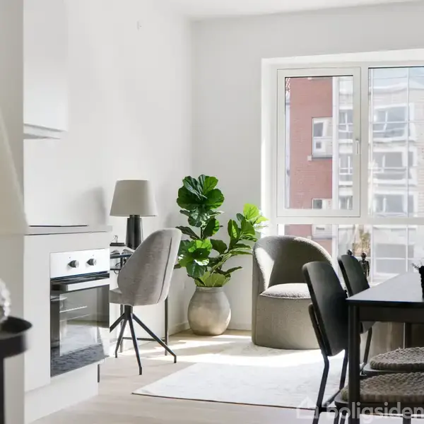 Spisebord med stole står i lys stue ved stort vindue, omgivet af moderne indretning, herunder lakeret gulv, indbygningsovn, stol ved skrivebord og grøn plante i hjørnet.
