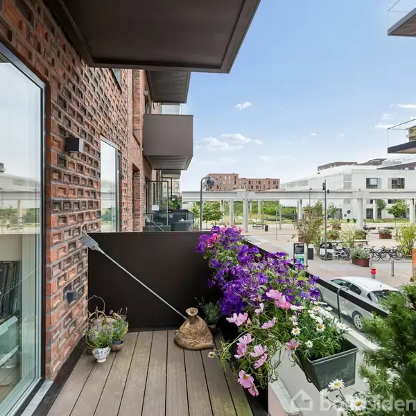Balkon med planter i blomsterkasser. Udsigt over moderne boligområde med parkerede biler og lejlighedsbygninger i baggrunden.
