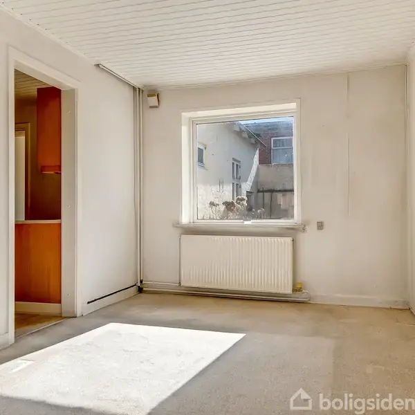 Et tomt rum med et vindue i midten af væggen, hvor sollys strømmer ind. På væggen under vinduet er der en radiator. Til venstre ses en åbning til et andet rum.