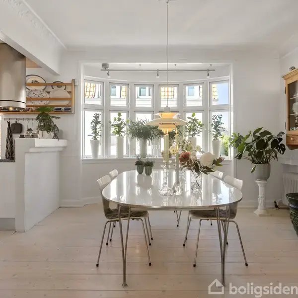 Et ovalt spisebord med seks stole står i et lyst rum. Bordet er dekoreret med blomster, og store vinduer giver naturligt lys. Planter og en træskænk omkranser rummet.
