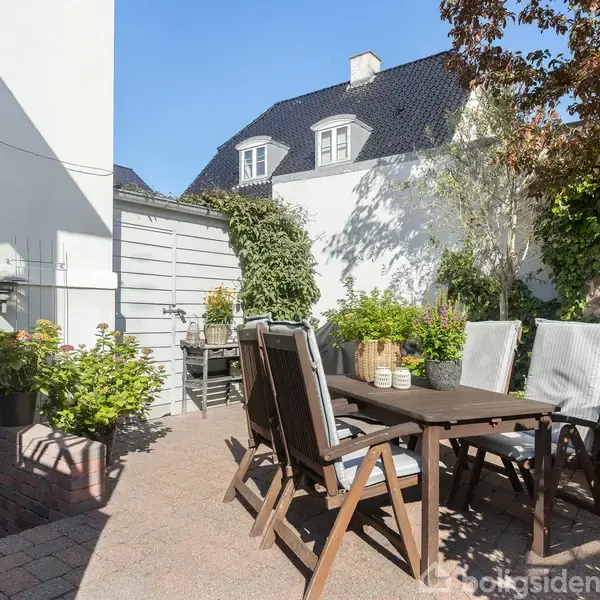 Terrassemøbler står på en solrig gårdhave med flisebelagt gulv og omgivet af planter og buske. I baggrunden ses en hvid bygning med sorte tagsten.
