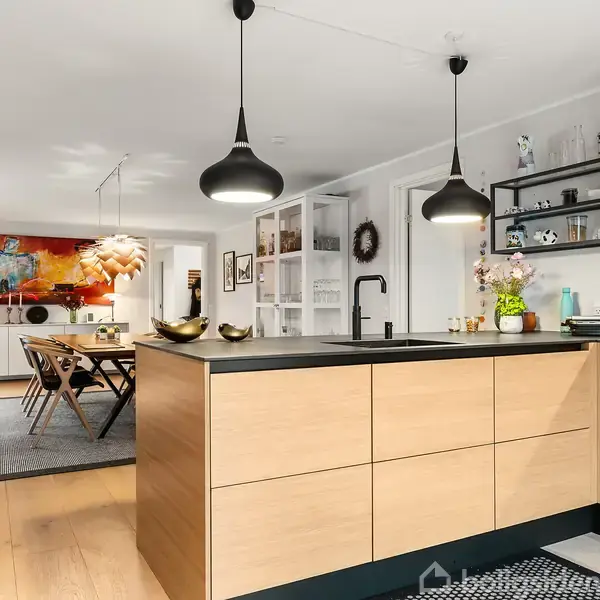 Køkkenø med vask og træskabe fremstår i et moderne køkken med sort bordplade. To pendellamper hænger over øen. I baggrunden ses spisebord og farverig kunst på væggen.