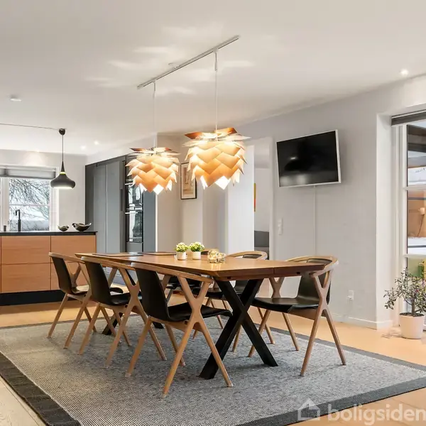 En spisebordgruppe med seks stole står på et tæppe. Over bordet hænger tre dekorative lamper. I baggrunden ses et moderne køkken og en vægmonteret fladskærm.