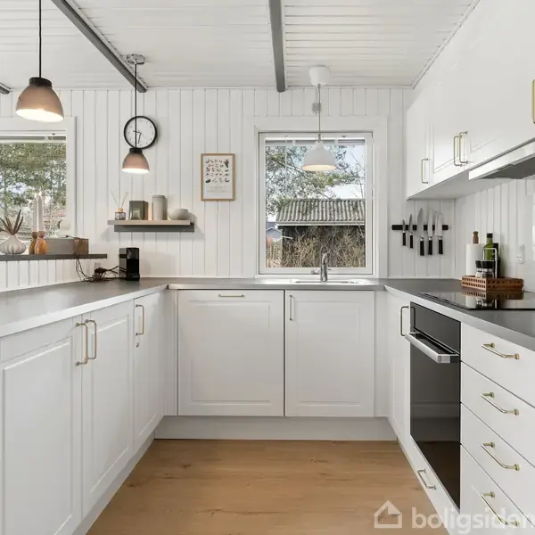 Hvidt køkken med sorte bordplader og moderne apparater, omfatter vægmonteret ur og hylde med krukker. Store vinduer giver udsigt til en have. Loftspendler belyser rummet.