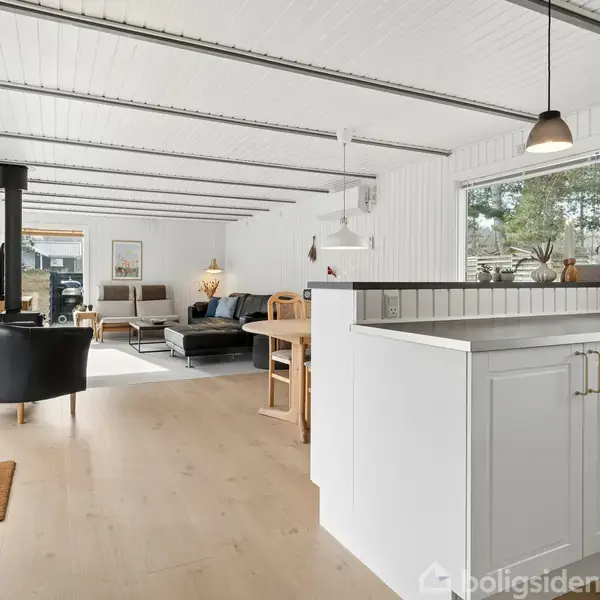 Køkken åbner ind til stue. Hvide skabe og mørke lamper. Stue har sort lænestol, hjørnesofa, brændeovn og spisebord. Store vinduer giver en lys og rummelig atmosfære.