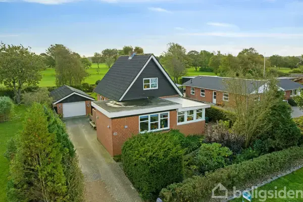 En murstensvilla med sort sadeltag står ved siden af en garage. En indkørsel og have omkranser huset. Baggrunden viser en grøn, skovklædt landskab.