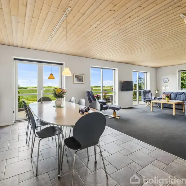 Stor stue med spisebord i forgrunden, omgivet af stole. Komfortable sofaer står i baggrunden. Flere vinduer viser en grøn udsigt, og rummet har lyse trælofter. Ingetekst synlig.
