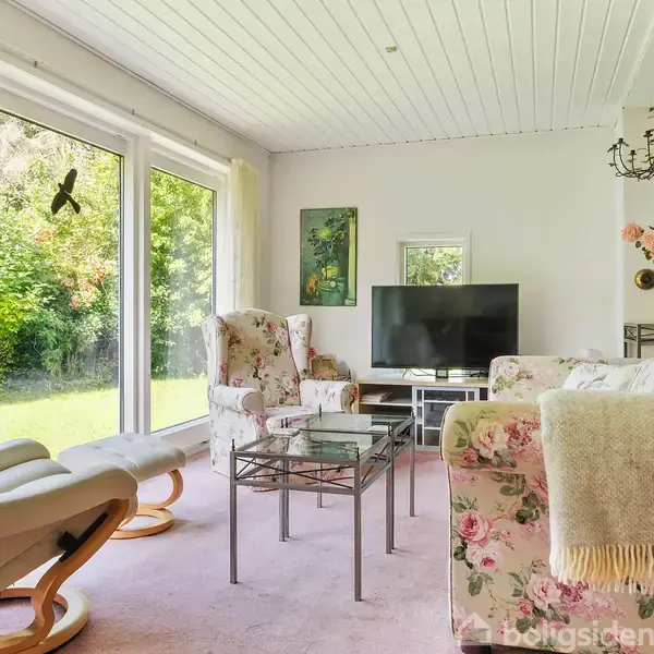 Stue med blomstermøbler og gyngestol, set mod et stort vindue med udsigt til grøn have. Et tv står overfor sofaen; loftet har en lysekrone. Uden tekst.