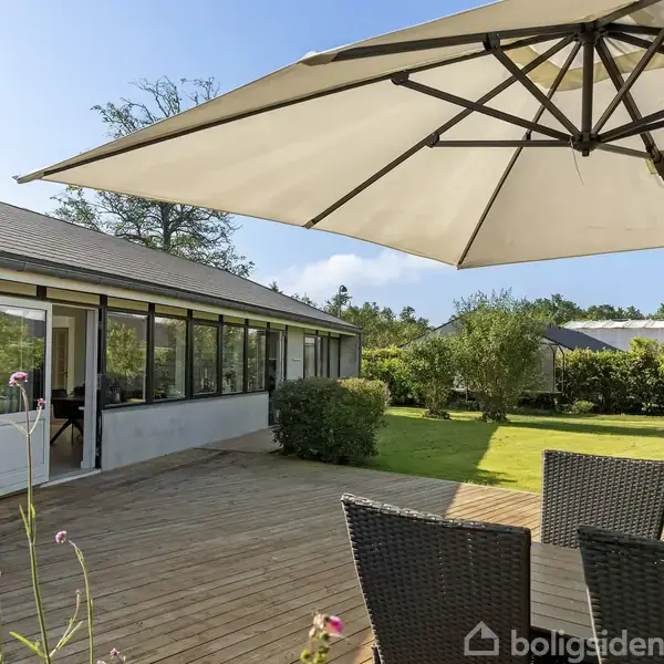 En terrasse med et stort hvidt parasol og sorte fletstole, placeret foran en bygning med store vinduer, omgivet af en grøn have og blå himmel.
