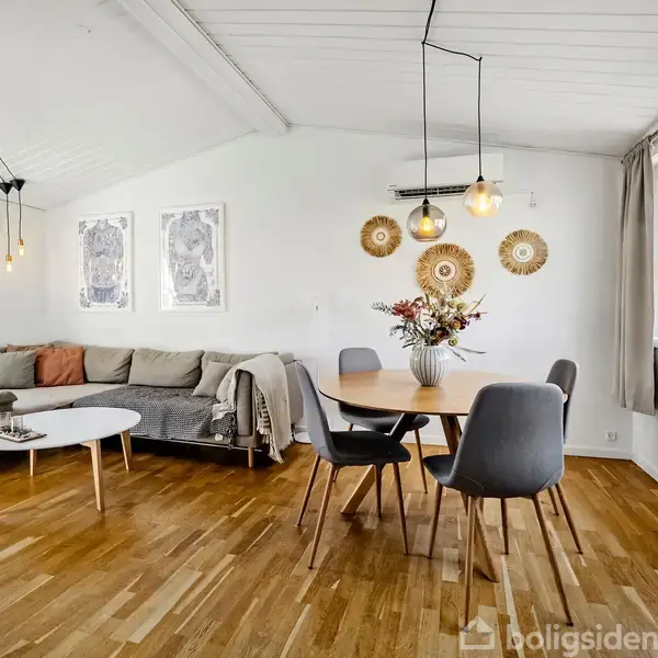 Sofa står i hjørnet med puder og plaid ved loungebord, lyse vægge. Rundt spisebord med fire stole og blomsteropsats. Store vinduer slipper lys ind. Moderne indretning med kunst på væggen.