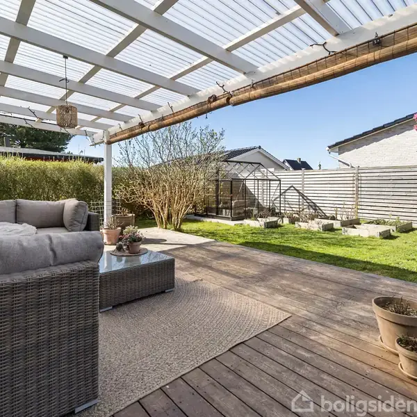 En udendørs veranda med en grå sofa under et halvtag af glas. Sofaen står på et trædæk med udsigt til en græsplæne og hegn. Planter i krukker er placeret rundt omkring.