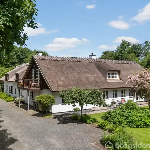 Traditionelt stråtækt hus står stille omgivet af en frodig have med træer og buske. I baggrunden ses en blå himmel med spredte hvide skyer.