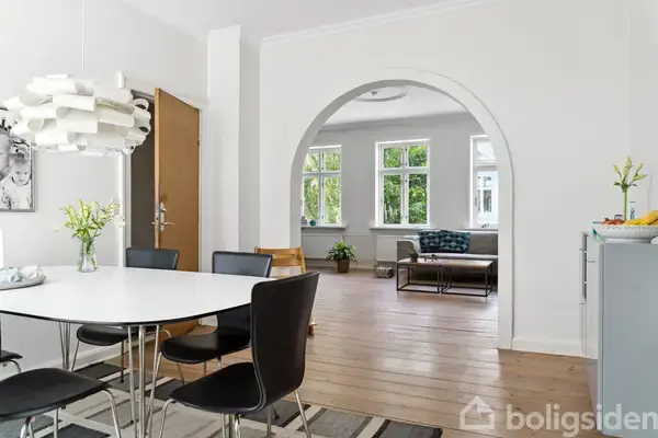 Et spisebord med sorte stole står i et lyst rum. Gennem en bue ser man en stue med vinduer og en sofa. Planter pynter rundt omkring.