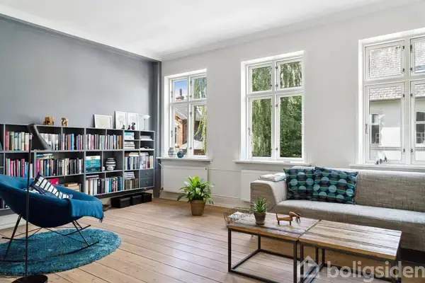 En stue med en grå sofa og et sofabord. En bogreol fyldt med bøger står ved siden af en blå lænestol. Store vinduer giver udsigt til en grøn have.