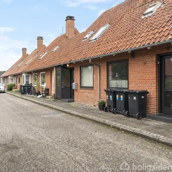 En række rødstensrækkehuse med skråt tag og sorte skraldespande foran ligger langs en smal, asfalteret vej. En hvid bil er parkeret længere nede ad vejen.