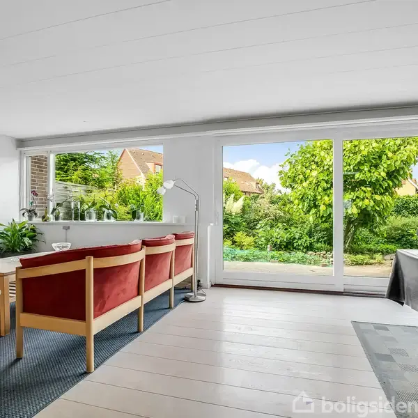 Stue med røde sofaer, et tv og et gulv-til-loft vindue, der åbner op til en grøn have med buske og træer. Hvidt loft og lyse trægulve.