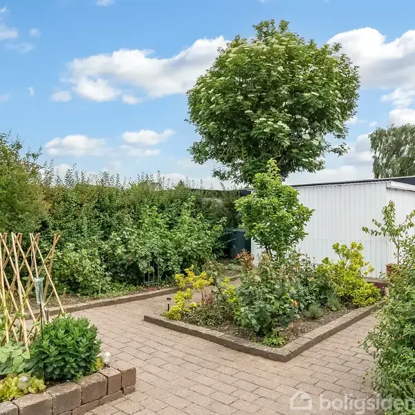 Have: En have med flisebelagt sti og hævede bede med planter. Kontekst: Omkranset af buske, med et stort træ og en hvid bygning i baggrunden under en blå himmel med skyer.