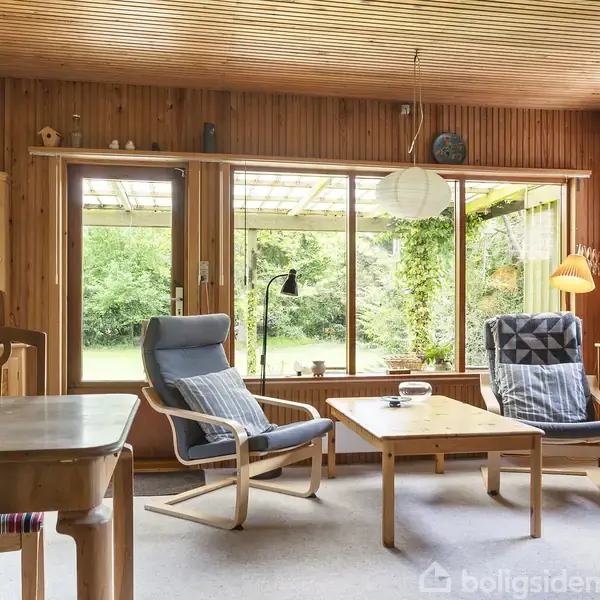 Stue med træpaneler, to polstrede lænestole ved et lavt træbord, store vinduer mod en grøn have. Til venstre ses spisebord og stole. En lampe hænger fra loftet.