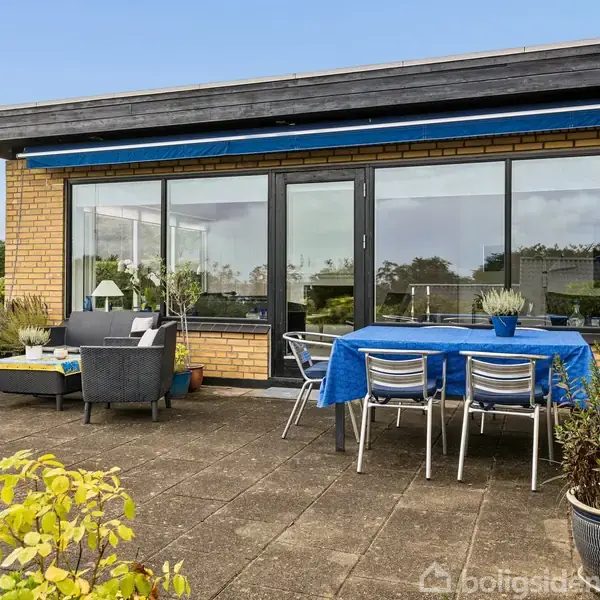 Terrassemøbler står på en flisebelagt terrasse foran et murstenshus med store vinduer. Et bord dækket med en blå dug er omgivet af stole, mens grønne planter omkranser området.