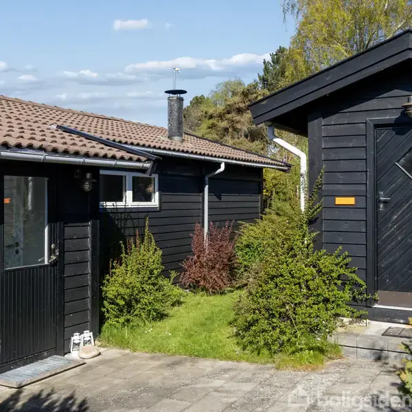 Hus med sort træfacade og rødt tegltag står i sollys. Grussti med omkransende buske fører til hoveddøren. Blå himmel og træer i baggrunden skaber en hyggelig stemning.