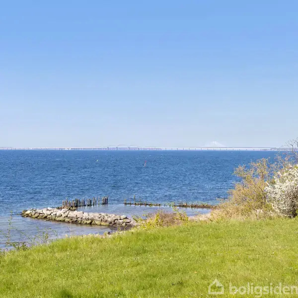 Bænk står stille i græsset ved havkanten, omgivet af frodig vegetation. Blå himmel og en rolig havudsigt med en lille stenmole fremhæver den rolige atmosfære.