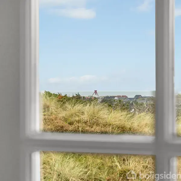 Et vinduesparti viser udsigt til en naturfyldt kyststrækning med græs og lave bygninger. I horisonten ses en blå himmel med få skyer, hvilket skaber en fredelig atmosfære.