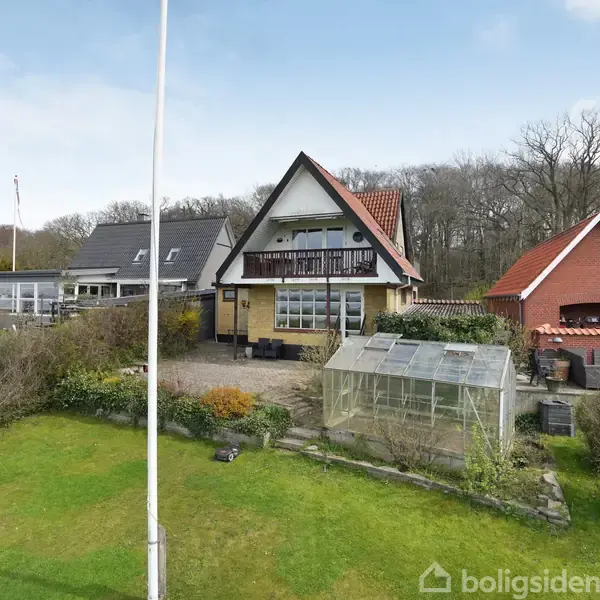 Et A-formet hus står stille i en velplejet have med en flagstang. Ved siden af huset er et drivhus, omkranset af buske. Skoven er i baggrunden af haven.