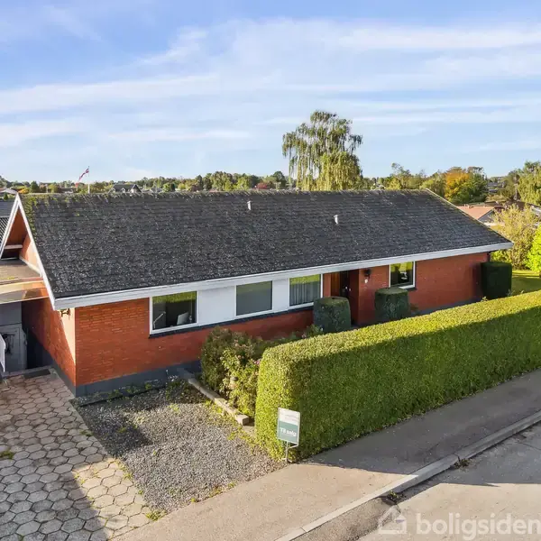 En ældre, rød murstensvilla med fladt tag står midt i et grønt villakvarter. En rød bil holder i carporten til venstre. Hækken omkranser haven og indkørslen.