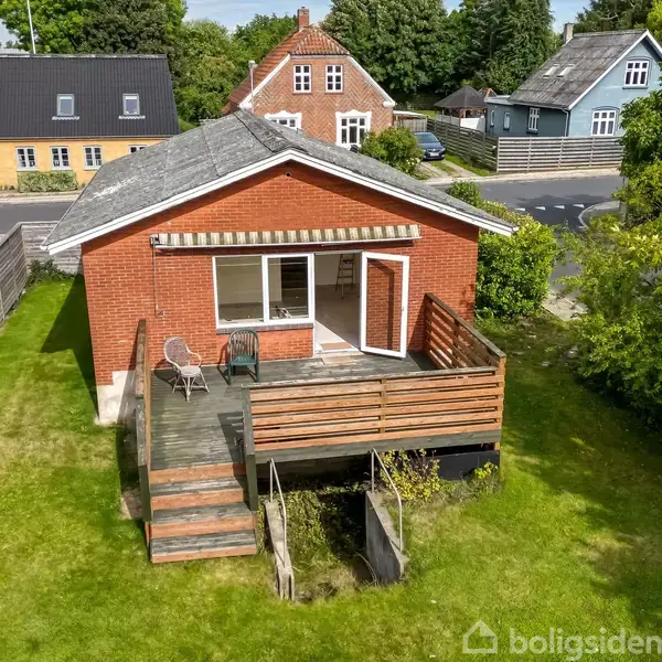 Rødt murstenshus med åben veranda, to stole og bord; beliggende i en villakvarter med grønne områder og flere huse i baggrunden på en solskinsdag.