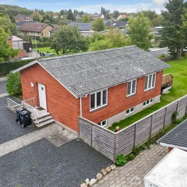 Rødt murstenshus har en grå tag og fem vinduer, står på en grusvej ved en grøn have med træer. Huset er omgivet af en træhegn og har en terrasse ved siden af.
