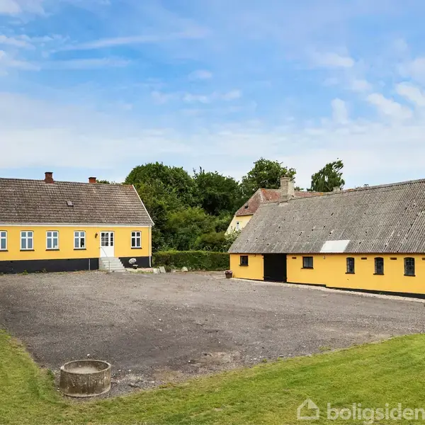 To gule, traditionelle bygninger står i en gårdsplads omgivet af grønt græs og træer under en blå himmel. En brønd er placeret i forgrunden til venstre.