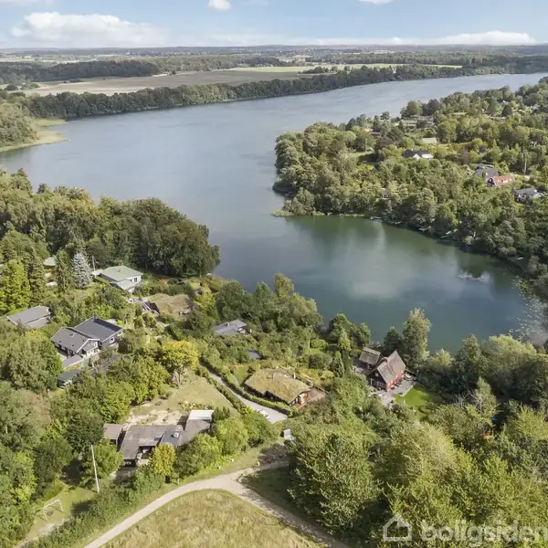 En sø omgivet af skov og spredte huse danner en naturskøn udsigt. Husene er beliggende i ujævnt terræn, og søen strækker sig gennem et frodigt landskab.