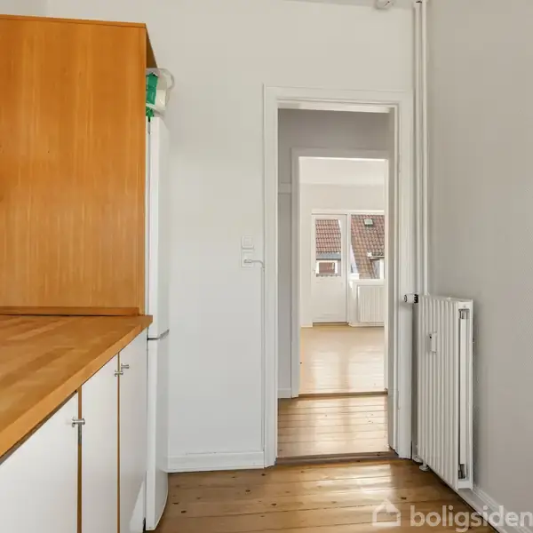 Et køkken med træbordplade og hvidt skab. Køkkenet fører gennem en dør til et lyst rum med trægulv og skråvinduer. Radiator på højre væg.