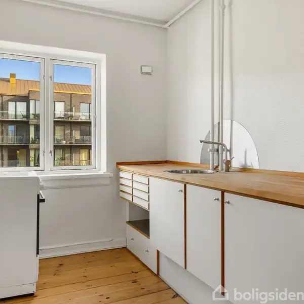 Køkken med hvide skabe og en vask, står i et rum med lyse trægulve. Et vindue viser en bygning udenfor. En komfur står ved siden af vinduet.