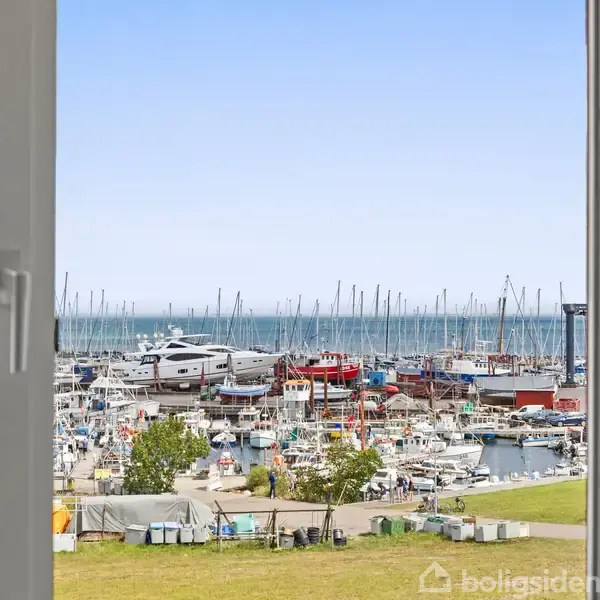 Åbent vindue giver udsigt til marina, hvor mange både ligger til kaj. I baggrunden ses åben hav og blå himmel. Foran marinaen er der en græsplæne med nogle træer.