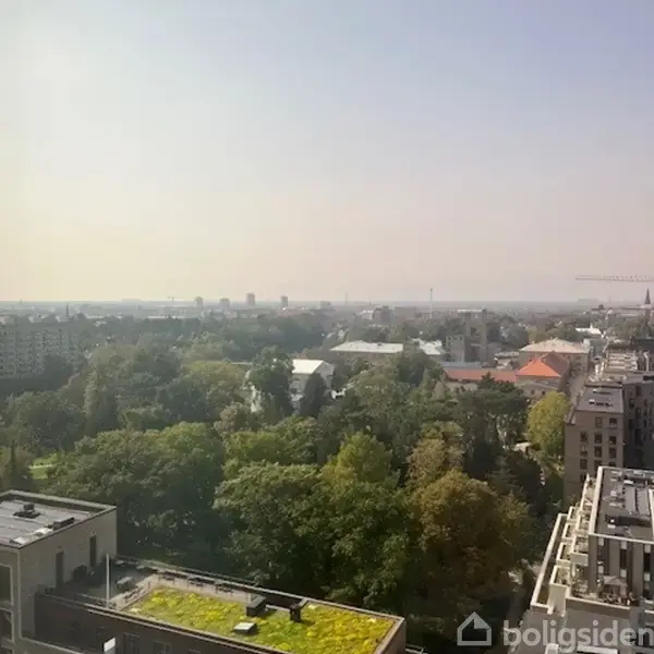 Udsigt over bylandskab om dagen med tæt bevoksede grønne træer i forgrunden. Bygninger og tårne strækker sig mod horisonten under en klar, lysende himmel.