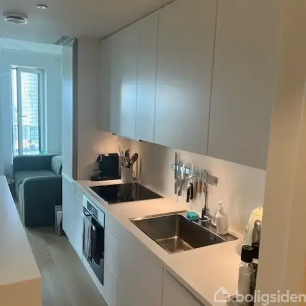 Et moderne køkken med hvide skabe over bordpladen; vask og komfur indbygget. Redskaber og rengøringsartikler ved vasken. Sofaområde og vindue i baggrunden.