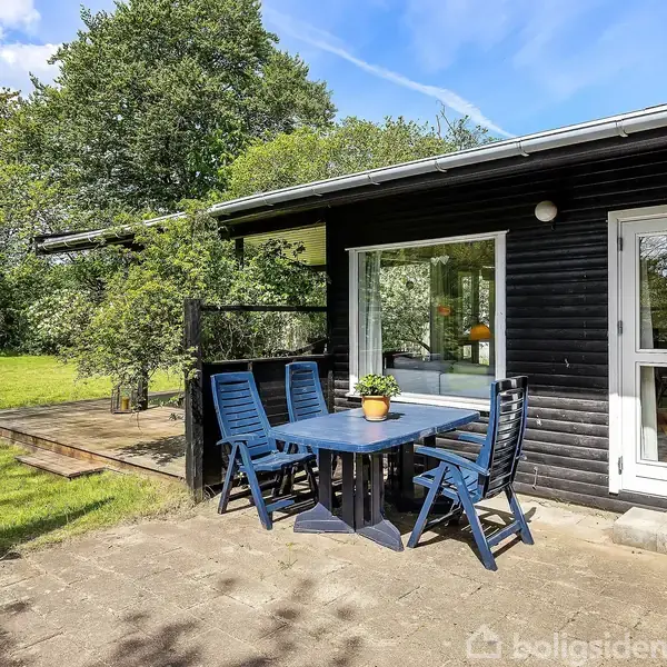 Sort træhus med terrasse, hvor et bord og fem blå stole står. En plante pryder bordet. Haven omkranser terrassen med græsplæner og træer; en grøn vandkande står nær.