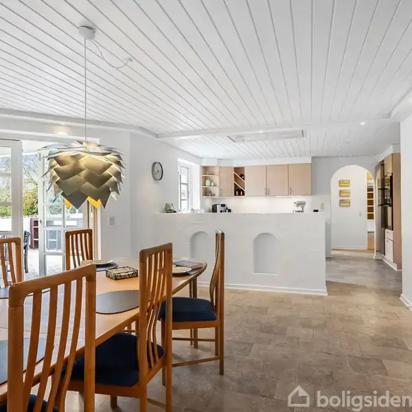Stue med spisebord omgivet af træstole og stor hængelampe. I baggrunden ses et hvidt køkken med åbne hylder. Store vinduer giver udsigt til terrasse og have.