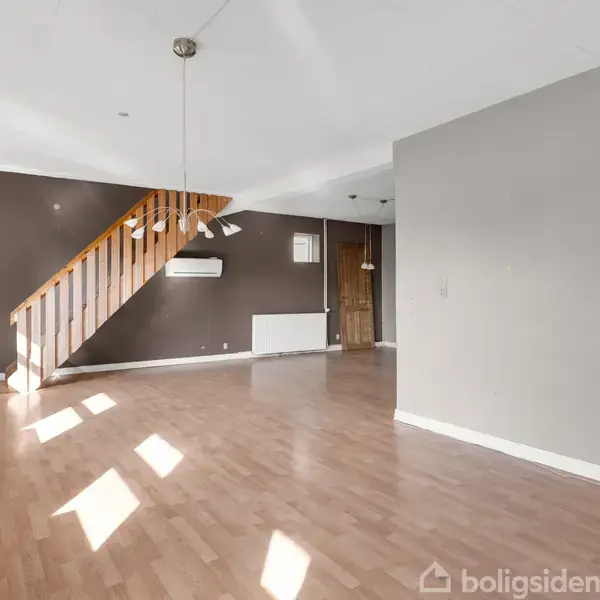 En stor, tom stue med trægulv og en trappe, der fører opad. Rummet er lyst med store vinduer og grå vægge. En dør fører til et køkken med hvide elementer.