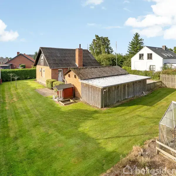 Rødt murstenshus med sort tag står midt i en stor græsplæne. Ved siden ligger en garage, og foran er et drivhus. Omgivet af grønne hække og træer.