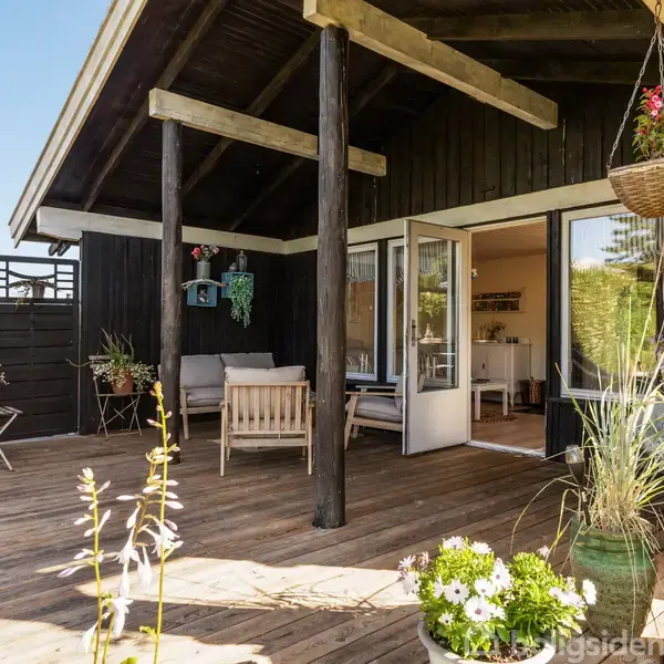 Et lyst, indendørs spiseområde med et hvidt bord og stole står foran åbne franske døre, der fører til en solrig terrasse og en frodig have med grønne buske.
