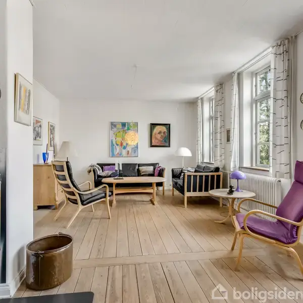 Brændeovn står i stue med flamme indeni, omgivet af en sofa, stole og et bord på trægulv. Væggene er dekoreret med malerier, og store vinduer slipper lys ind.
