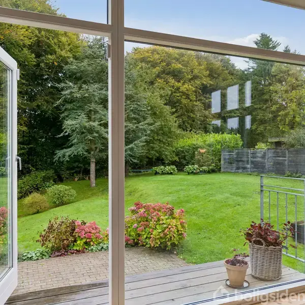 En glasdør står åben og fører ud til en frodig have med grønne buske og træer. En terrasse med blomsterkrukker er i forgrunden, omgivet af græsplæne.