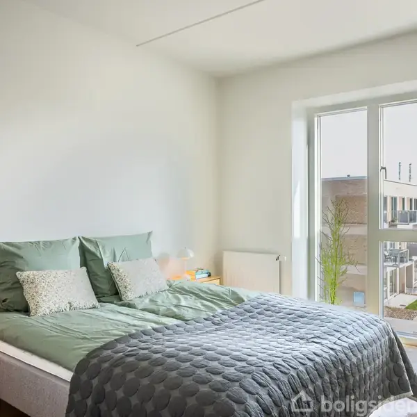 En seng med grønne puder og et gråt sengetæppe står ved siden af et lille natbord med en lampe, i et soveværelse med store vinduer og udsigt til en moderne boligblok.