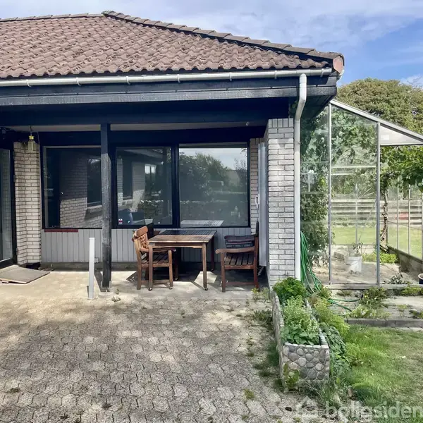 En terrasse med to træstole og et bord står foran et hus med mursten og mørkt tag. Til højre ses et drivhus omgivet af græs og planter.