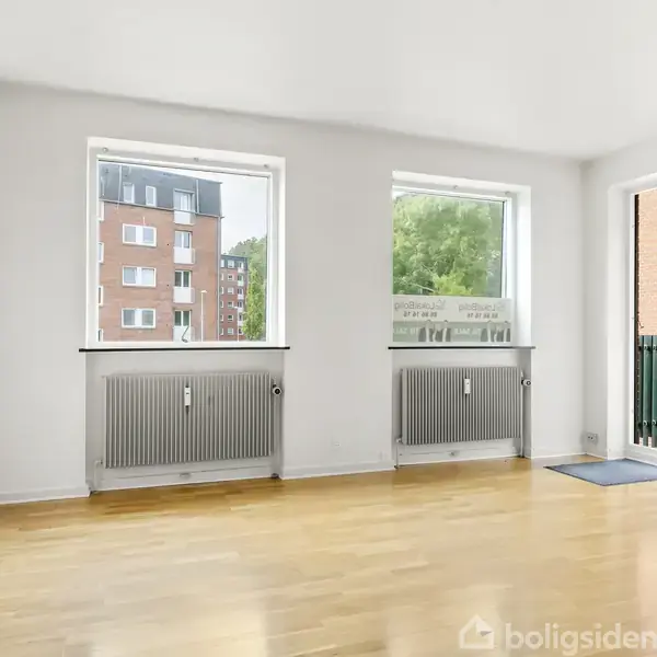 Et lyst, tomt rum med trægulv har to vinduer og en altandør, der fører ud til en lille balkon. Radiatorer er monteret under vinduerne. Udsigt til bybygninger og grønne områder.
