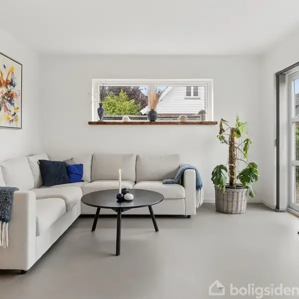 En hvid sofa står i en lys stue med kunst på væggen, et rundt sofabord foran, og grønne planter ved siden af. Store vinduer giver udsigt til en have.