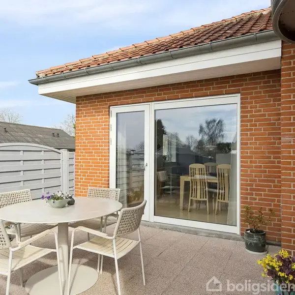 Rundt havebord med fire stole står på terrasse af fliser. Bagved er en murstensbygning med vindue og dør. Grå hegn omgiver området, og der er blomsterdekorationer.
