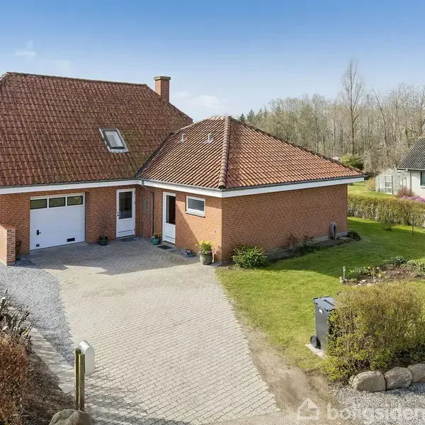 Murstenshus med rødt tegltag står på en velplejet grund med en stenterrasse og grøn græsplæne. Omgivet af hække og haveanlæg, huset har en garage og flere vinduer.